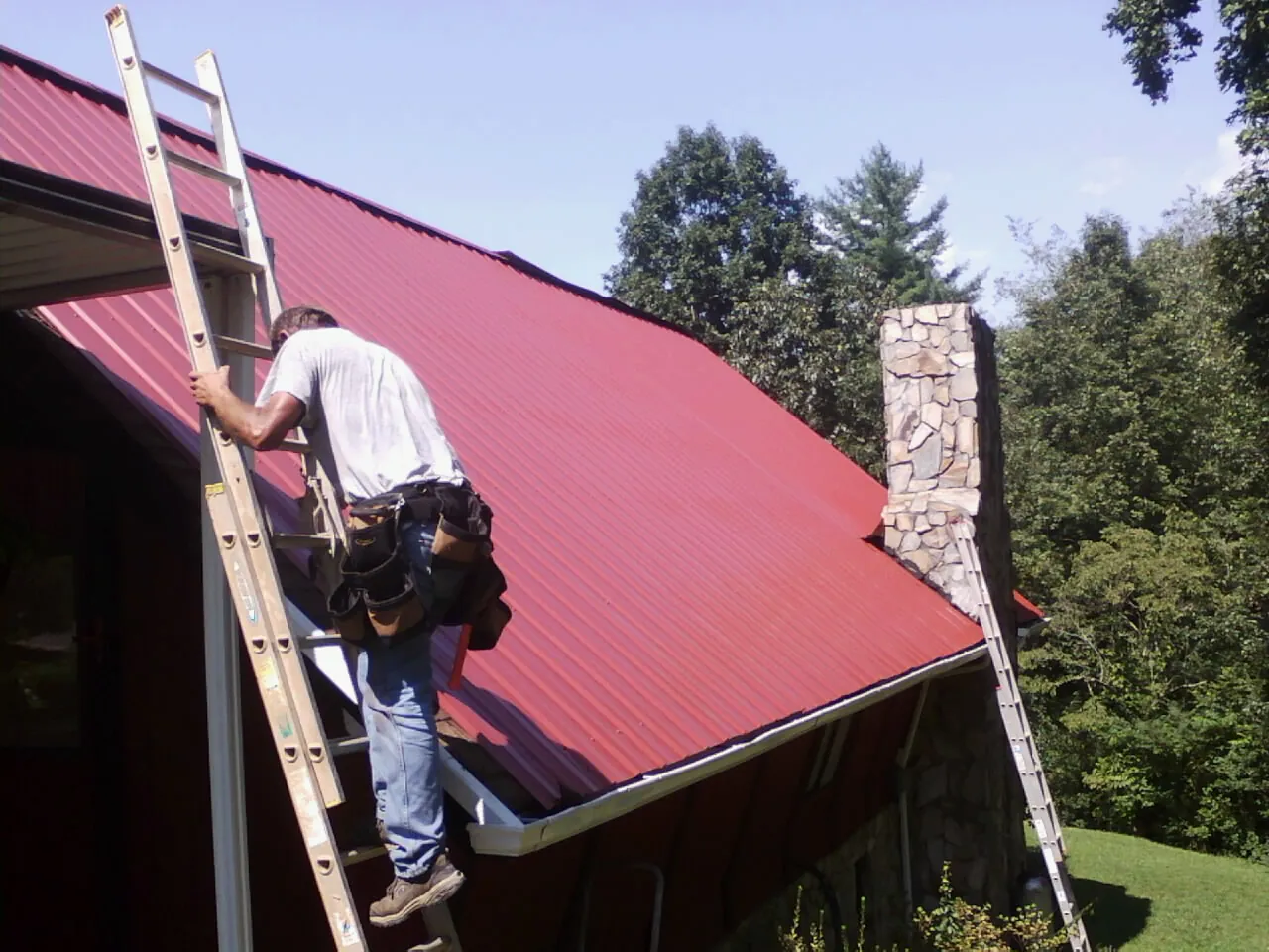 Roofer on a ladder beside a red metal roof and stone chimney — Soffit & Fascia Repair by Roofing Brothers in Madison Heights